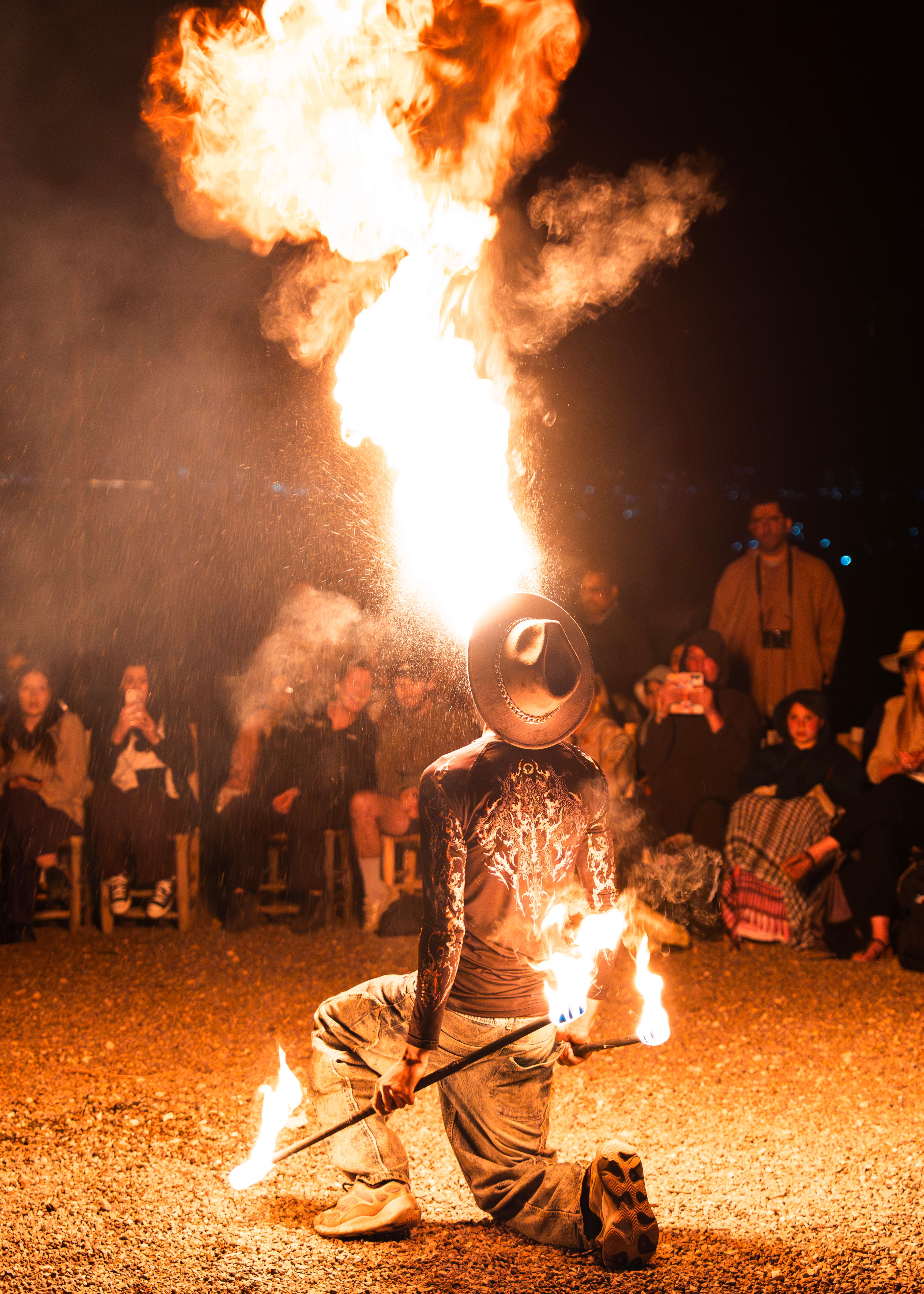 Fire performer kneeling and breathing a tall flame for guests seated around the desert dinner camp